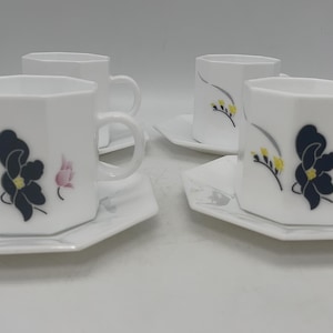 May include: Four white ceramic teacups and saucers. Each cup has a unique octagonal shape and a handle. The cups and saucers are decorated with a black flower design, yellow accents, and pink flowers.