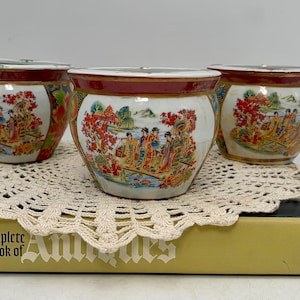 May include: Three small, round ceramic pots with a red rim and a detailed Asian-inspired scene. The pots are white with colorful illustrations of people, trees, and landscapes. They sit on a cream-colored doily atop a book.