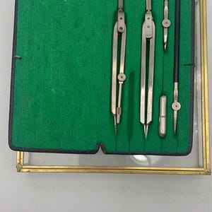 May include: A collection of vintage drawing instruments, including compasses and dividers, presented in a green felt-lined case with a gold-coloured frame. The tools are silver and appear to be well-preserved, indicating careful upkeep.