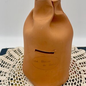 May include: A terracotta piggy bank shaped like a bag with the text "Il Sacco di Soldi" on the front.
