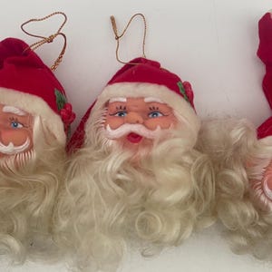 May include: Three vintage Santa Claus head ornaments with white beards and red velvet hats. The ornaments are made of plastic and have a festive Christmas theme.