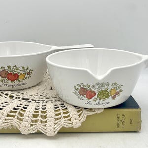 May include: Two white ceramic sauce pots with handles. The pots feature a floral design with the words "La Magdelina" and "La Sauge" printed on them.