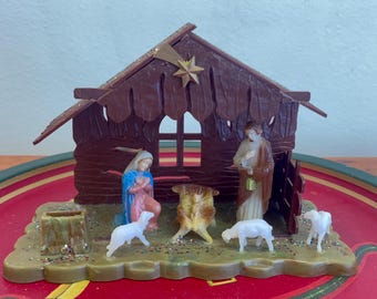Pode incluir: Um presépio vintage de plástico com um estábulo de madeira marrom, uma estrela no topo e figuras de Maria, José e um bebê Jesus. Há também duas ovelhas de plástico brancas na cena.