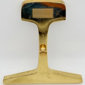 May include: A polished gold-tone metal paperweight shaped like a railroad track cross-section. The top section features a rectangular plaque engraved with the text "CONRAIL". A small, cylindrical knob is attached to the centre of the vertical stem.