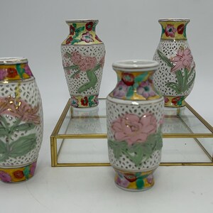 Puede incluir: Cuatro pequeños jarrones de porcelana blanca con intrincados diseños florales. Los jarrones están decorados con flores rosas, amarillas y verdes y tienen un delicado patrón de encaje. Se exhiben en un soporte de vidrio transparente con un borde dorado.