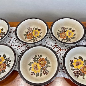 May include: A set of seven round ceramic bowls with a cream-colored interior and a dark brown rim. Each bowl features a central floral pattern consisting of yellow and orange flowers with dark green leaves and small brown accents.