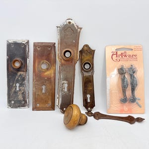 Peut inclure: Divers éléments de quincaillerie de porte anciens, comprenant des plaques rectangulaires avec des trous de serrure, un bouton de porte en bois et des poignées de tiroir en forme d'hippocampe. Les pièces métalliques présentent des signes d'usure et de rouille.