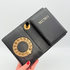 Puede incluir: Libreta de direcciones de polipiel negra con un dial giratorio dorado y un pequeño bloc de notas. El bloc de notas está grabado con la palabra "MEMO" en dorado. La libreta de direcciones está abierta, revelando el dial y el bloc.