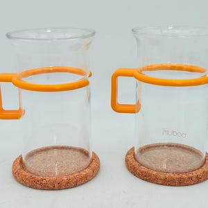 May include: Two clear glass mugs with orange handles and rims, resting on round cork coasters. The mugs have a cylindrical shape with a slightly flared top. The brand name "muboa" is printed on the side of one mug.