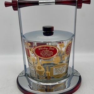 May include: Vintage chrome and glass ice bucket with a red handle and base. The glass bucket features gold lettering and illustrations, including a recipe for a "Whiskey Sour" cocktail. The bucket is approximately 7 inches tall.