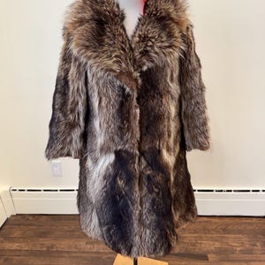 May include: A full-length fur coat in shades of brown, gray, and black, displayed on a mannequin. The coat features a large, plush collar and a long, flowing design. A red fabric is visible at the neck.