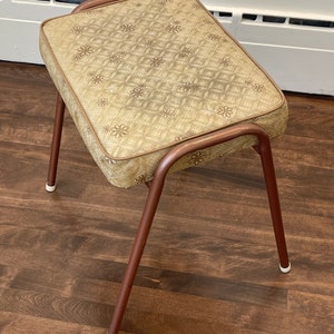 May include: A small, brown, upholstered stool with a floral pattern on the seat. The stool has a metal frame with four legs and white plastic feet.