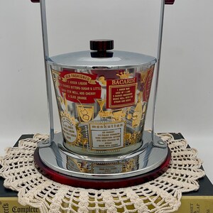 May include: Vintage chrome and glass ice bucket with a red handle and base. The glass is decorated with gold lettering and cocktail recipes, including "Old Fashioned" and "Bacardi". A stylish barware item.
