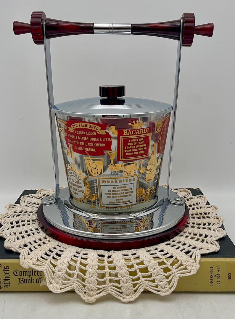 May include: Vintage chrome and glass ice bucket with a red handle and base. The glass is decorated with gold lettering and cocktail recipes, including "Old Fashioned" and "Bacardi". A stylish barware item.