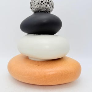 May include: A sculpture composed of stacked elements: a light orange wooden base, a white stone, a black stone, and a silver sphere with a textured surface. The background is plain white.