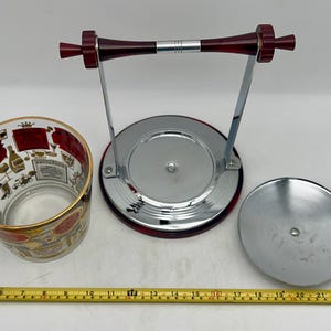 May include: A vintage ice bucket set with a clear glass bucket featuring a gold rim and red and gold illustrations. The set includes a chrome stand with a red handle and a lid. The set is designed for serving beverages.