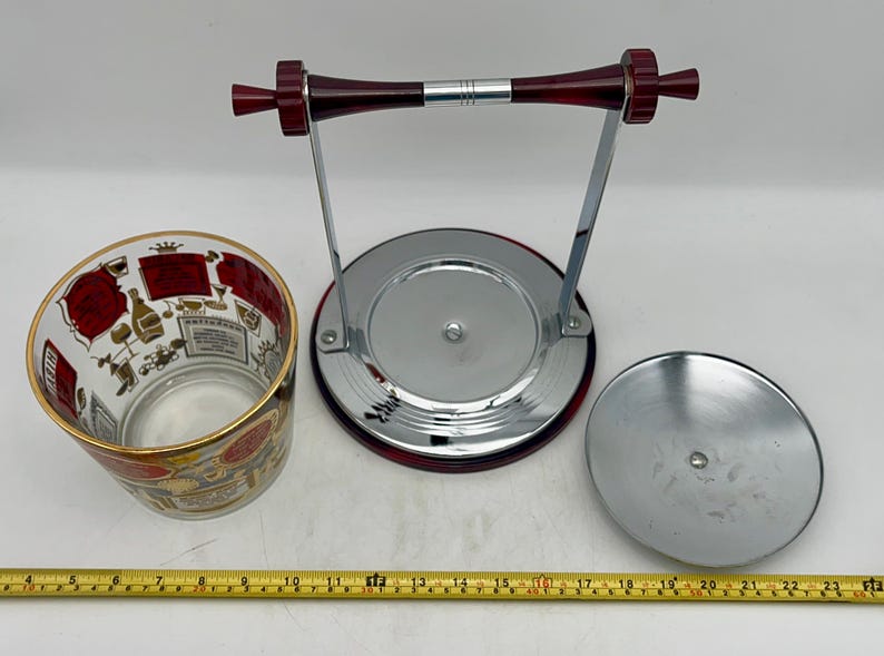 May include: A vintage ice bucket set with a clear glass bucket featuring a gold rim and red and gold illustrations. The set includes a chrome stand with a red handle and a lid. The set is designed for serving beverages.