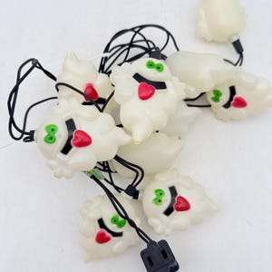 May include: A string of decorative lights featuring multiple off-white ghost-shaped covers. Each ghost has a black mouth, a red heart-shaped tongue, and bright green eyes. The figures are connected by a black electrical cord with a black plug.