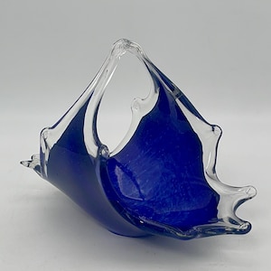 May include: A glass basket with a deep blue interior and a clear, arched handle. The basket has an unusual, asymmetrical shape with pointed edges. The glass has a textured surface. The piece is likely handmade.