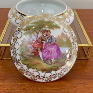 May include: A white porcelain trinket box with a lid featuring a romantic scene of two figures in a garden, with gold filigree accents. The box is open, revealing a white interior. The lid is decorated with a painting of a couple in a garden setting.