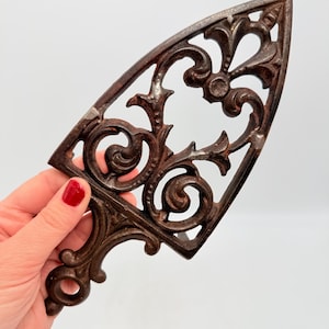 Puede incluir: Trivet de hierro fundido ornamentado con acabado marrón oscuro. El salvamanteles presenta un diseño decorativo calado con patrones de volutas y un asa. Accesorio de cocina vintage.
