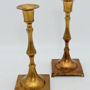 Puede incluir: Dos candeleros de latón vintage. Cada candelero tiene una base cuadrada, un tallo delgado con elementos decorativos y una parte superior circular para sostener una vela. El latón tiene un tono dorado cálido y el diseño es clásico y elegante.