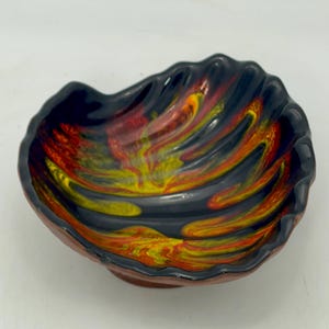Peut inclure: Un bol en céramique au design unique. L'intérieur présente un motif tourbillonnant de rouge, de jaune et d'orange sur un fond noir. Le bord est noir et l'extérieur est de couleur brun rougeâtre.