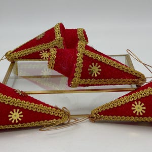 May include: Four red velvet Christmas decorations with gold trim and sequins. The decorations are shaped like cones and have a gold string for hanging.