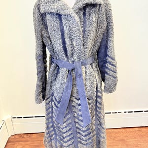 May include: A grey faux fur coat with a wide collar and a matching belt. The coat features a chevron pattern on the lower half and blue suede accents. The coat is displayed on a mannequin.