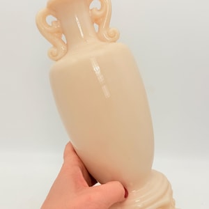 以下が含まれることがあります： 桃色の装飾的な花瓶で、蓋と2つの装飾的なハンドルが付いています。 花瓶は丸みを帯びたボディで、巻き貝の足が付いた装飾的なベースに置かれています。 花瓶は人が持っています。