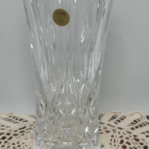 May include: Clear Capri crystal vase with a diamond-cut pattern. The vase has a cylindrical shape with a flared top. A gold sticker on the front reads "CAPRI CRYSTAL OVER 24% PBO". The vase is sitting on a cream-colored, open-weave doily.