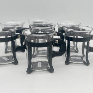 Peut inclure: Six tasses en verre transparent avec des poignées et des bases en métal noir. Les tasses sont disposées en rangée sur une surface blanche.