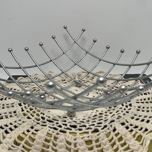May include: A silver-colored metal fruit basket with a geometric design. The basket is made of curved wires with small ball-shaped accents at the ends. It sits atop a cream-colored, crocheted doily and a vintage book.