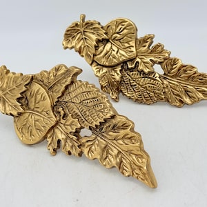 Peut inclure: Deux objets décoratifs dorés en forme de feuilles. Chaque objet présente des motifs de feuilles détaillés, notamment des érables et d'autres types de feuilles. Les objets sont probablement en métal et sont présentés sur un fond blanc.