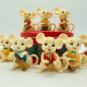 Op de afbeelding: Een verzameling vintage, crèmekleurige muizenbeeldjes, elk met een muziekinstrument. De muizen dragen kleurrijke vesten en vlinderdassen, met instrumenten als saxofoons, drums en gitaren. Ze zijn gerangschikt rond een rode blikken doos.