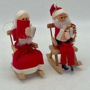 May include: A pair of vintage Christmas decorations featuring Santa Claus and Mrs. Claus seated in rocking chairs. Santa is dressed in a red suit with white trim and a white beard. Mrs. Claus is wearing a red dress with white trim and a white shawl. Both decorations have a white felt face with painted features and glasses.