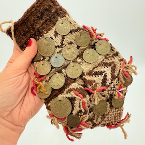 Puede incluir: Una bolsa de punto marrón y beige adornada con numerosas monedas antiguas. La bolsa presenta un patrón geométrico y está adornada con flecos rojos y amarillos. Las monedas están unidas a la bolsa.