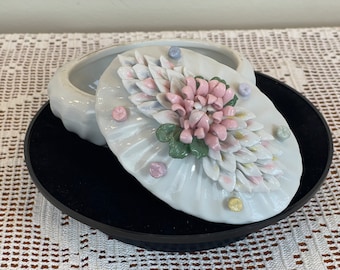 May include: White porcelain trinket box with a floral design. The lid features a pink and white flower with green leaves and white petals. The box is decorated with small, colorful dots.