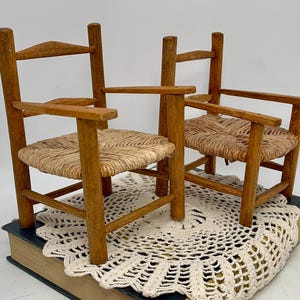 May include: Two small wooden chairs with woven seats. The chairs have armrests and a ladder-back design. The chairs are light brown and sit on a cream-colored doily. The chairs are a charming addition to any dollhouse or miniature collection.