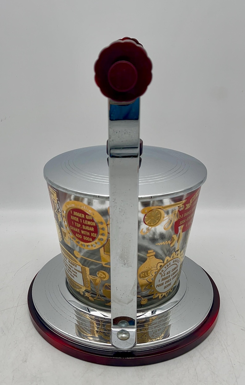 May include: Vintage cocktail shaker with a red and chrome finish. The glass cup features gold and red graphics with text. The shaker has a red handle and a chrome lever. The base is red and chrome.
