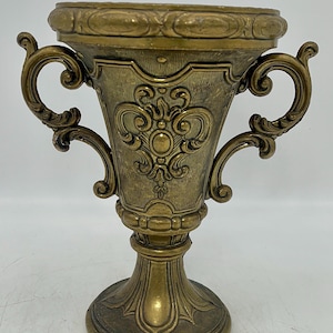 May include: An ornate antique brass-coloured chalice with two curved handles. The chalice features detailed relief patterns and a wide rim. The base is a pedestal design, adding to its vintage aesthetic.