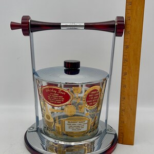 May include: Vintage cocktail shaker with a chrome and red base, handle, and accents. The glass container features gold lettering and cocktail recipes for Whiskey Sour and Bloody Mary. A wooden ruler is next to the shaker.