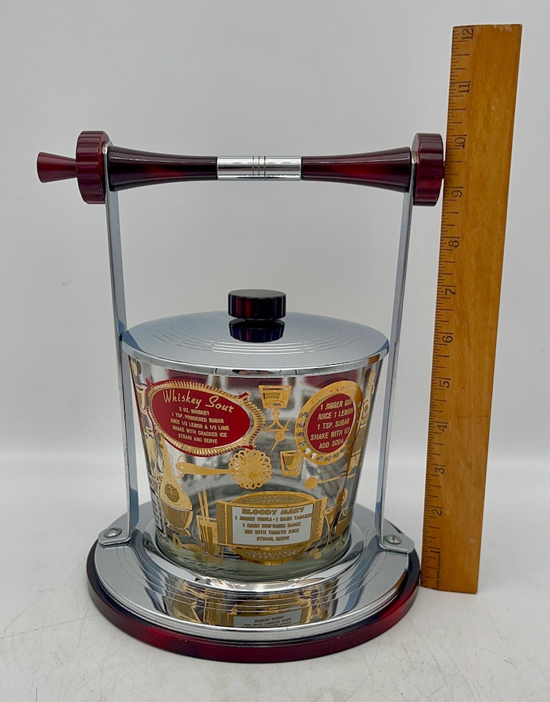 May include: Vintage cocktail shaker with a chrome and red base, handle, and accents. The glass container features gold lettering and cocktail recipes for Whiskey Sour and Bloody Mary. A wooden ruler is next to the shaker.