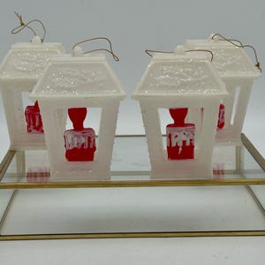 May include: Four white plastic Christmas decorations shaped like lanterns with red candle-shaped accents. The decorations are displayed on a glass and gold stand.