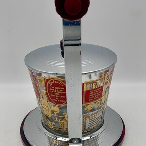 May include: Vintage cocktail shaker with a silver lid and base, and a clear glass body. The body features gold lettering and illustrations of cocktail recipes. The handle and top are red.