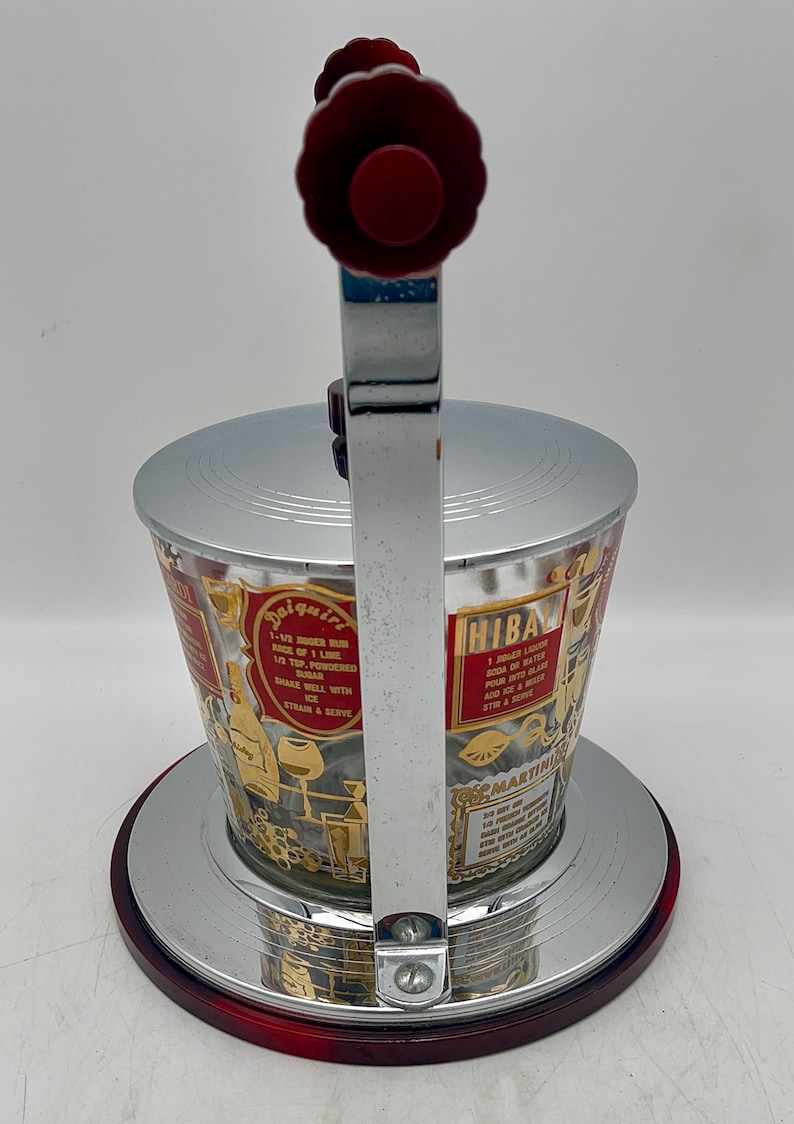 May include: Vintage cocktail shaker with a silver lid and base, and a clear glass body. The body features gold lettering and illustrations of cocktail recipes. The handle and top are red.