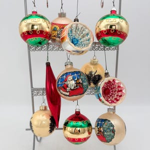 May include: A collection of eleven glass Christmas ornaments hanging on a gray metal display stand. The ornaments include spherical, teardrop, and indented designs featuring gold, red, and green colors, with painted scenes of Santa Claus.