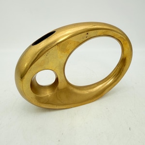 Puede incluir: Un jarrón de metal dorado pulido con una forma asimétrica, redondeada y hueca. El objeto presenta una superficie lisa y continua con una gran abertura central y una abertura circular más pequeña, rematada por una abertura estrecha y alargada.