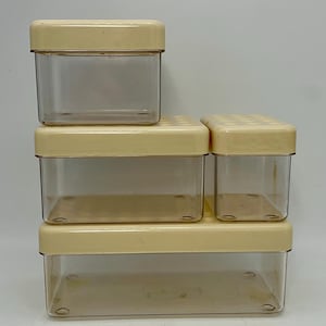 Puede incluir: Una pila de cuatro recipientes de almacenamiento de plástico transparente con tapas de color crema. Los recipientes son rectangulares y de diferentes tamaños, siendo el más grande el de abajo. Las tapas tienen una superficie ligeramente texturizada.