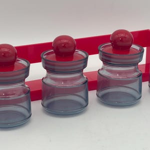 May include: Four clear glass spice jars with red lids on a red shelf. The jars are filled with spices.